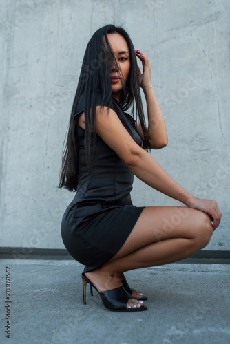 Obraz na plátne Side profile of a beautiful Asian woman crouching in a short black silk dress and high heels against a concrete wall