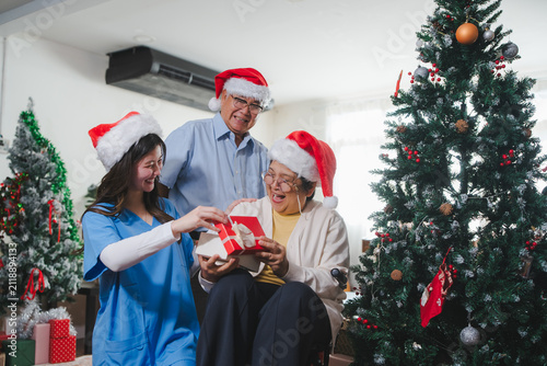 Joyful senior woman laughing while opening Christmas gift with husband and caregiver. Excellent for family retirement health insurance, inclusive nursing home, and festive corporate ads.