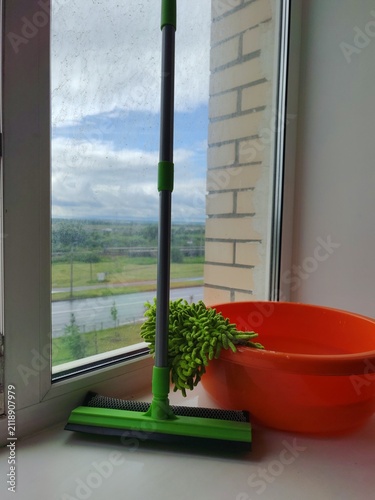 Window cleaning supplies with water bucket,squeegee, green sponge and cloth beside a dirty window in a home interior.