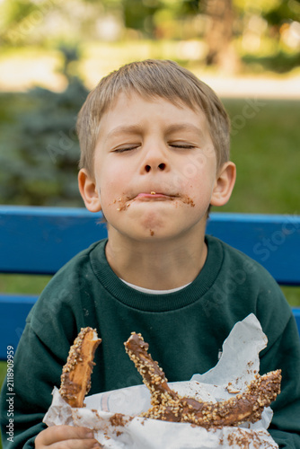 A cute little boy with a chocolate-stained face sitting on a blue bench outdoors, blissfully eating a delicious chocolate-covered pretzel with nuts with his eyes closed.