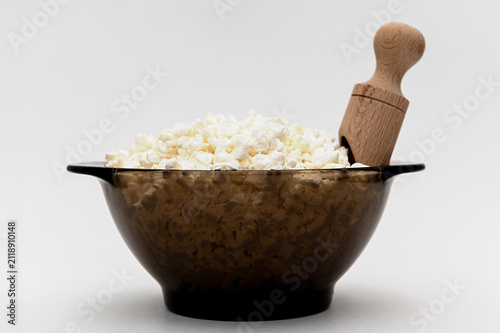 White crumbly cottage cheese with a wooden scoop in a black glass bowl on a white background.