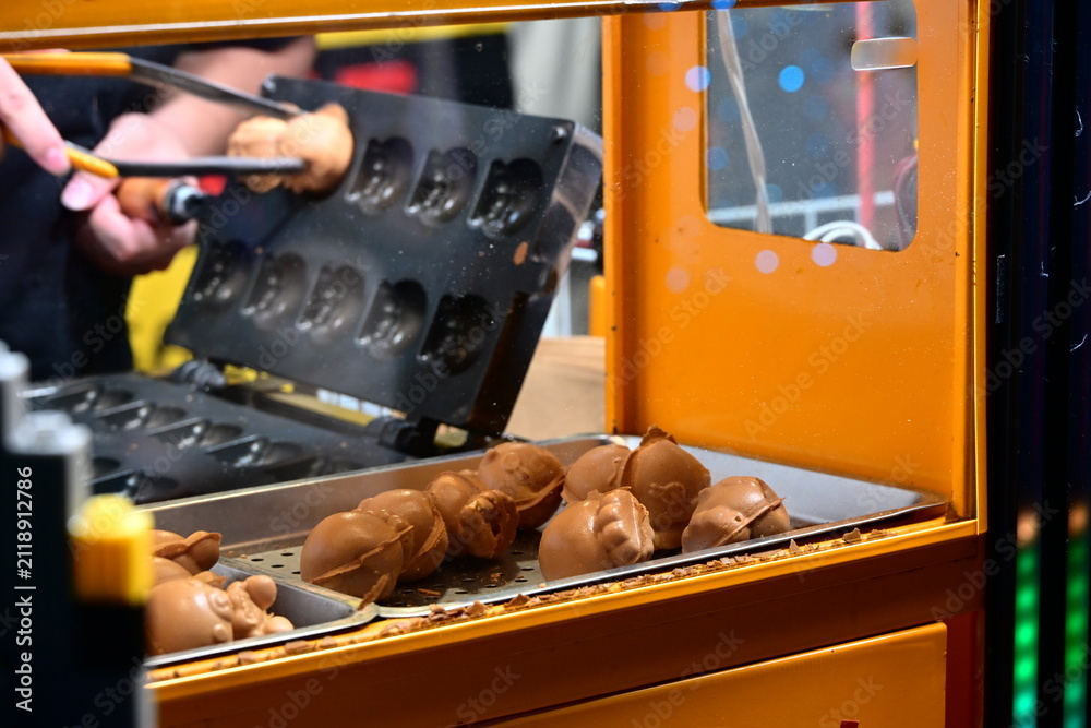 Fototapeta premium New Taipei City, Taiwan - Nov 16, 2025: Hands lift golden egg cakes from an iron mold in Banqiao District, steam rising beside a yellow street food stall.