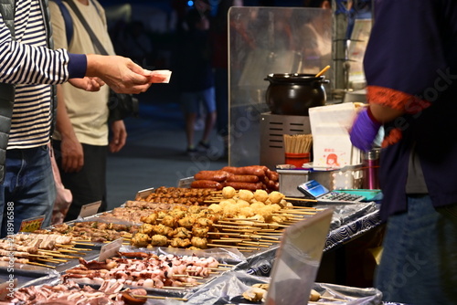 Canvastavla New Taipei City, Taiwan - Nov 16, 2025: Customer hand reaching for grilled skewers as vendor assists at night market stall, warm tungsten light on rows of meat skewers, Banqiao District night