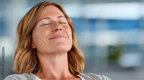 A middle-aged woman with light brown wavy short hair tilts her head back, eyes closed with a relaxed expression, set against a blurred cool-toned indoor office background.

