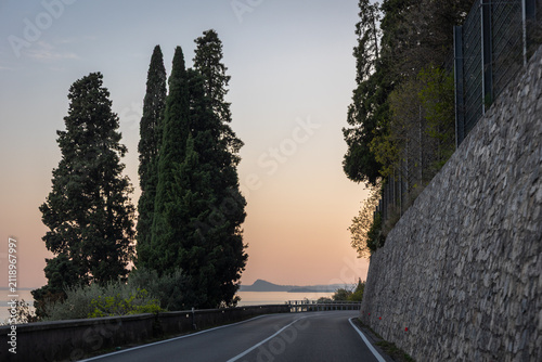 Scenic lakeside road lined with cypress trees at sunset on Lake Garda, Italy