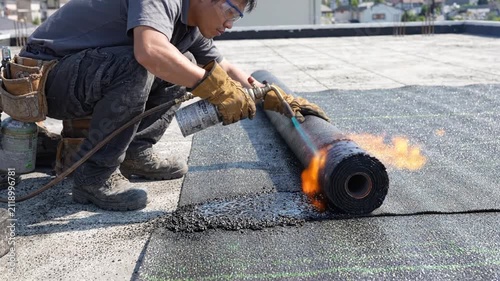 Roofer applying bitumen roll with torch on flat roof construction