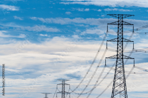 High voltage electricity transmission towers and power lines beneath a blue cloudy sky. Modern electrical infrastructure, utility grid reliability, energy distribution and sustainable power networks.
