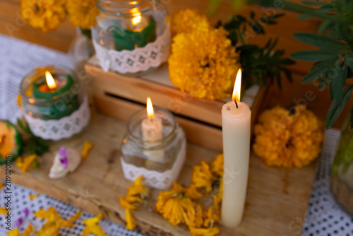 Close view of lit glass jar candles, taper flame, bright orange marigold flowers, green foliage, and rustic wood surface forming heartfelt memorial setting with warm soft light, with day, dead