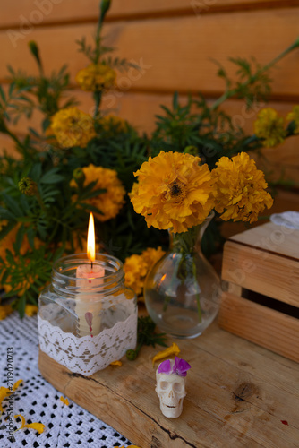 Closeup of bright orange marigold flowers in glass vases beside lace wrapped candle burning inside jar and small white decorative skull with purple petal, arranged on rustic wooden table