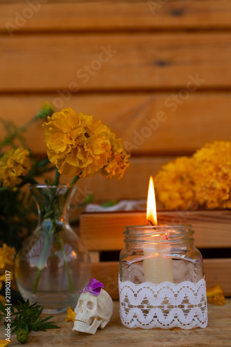 Closeup of bright orange marigold blossoms beside small decorative skull and burning candle in glass jar wrapped with lace on wooden background, warm soft light and shallow focus, with day, dead