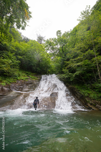 Canyoneer standing at the base of a cascading waterfall surrounded by lush green forest in Japan