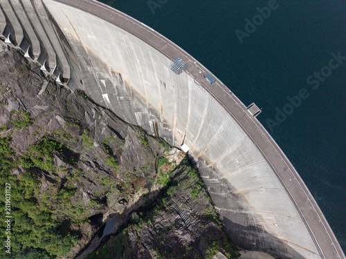 Aerial view of the massive concrete wall of the Contra Dam of Verzasca Hydroelectric Power Station nestled against steep rocky cliffs and deep water in Tenero-Contra, Ticino, Switzerland.