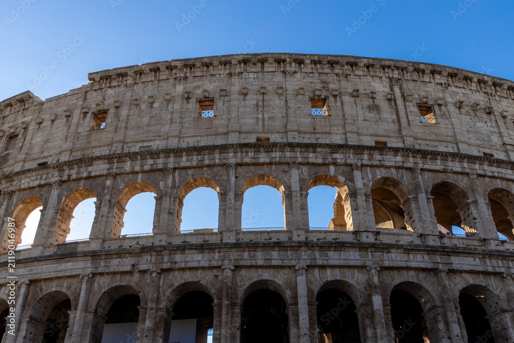 Obraz premium Famous Coliseum in Rome. Colosseum, or Coliseum. Part of the wall of an antique stone structure with arches. Italy, Europe. Famous touristic place in the world, Europe. Beautiful blue sky background.