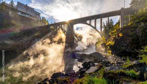 Majestic bridge over waterfall with sunburst, lush greenery, and mist.