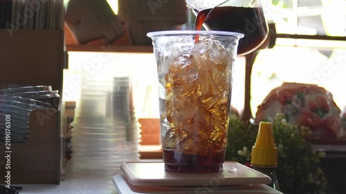 4K Pouring fresh brewed drip black coffee over ice cubes in plastic cup at cozy cafe counter, Concept for iced coffee barista craftsmanship summer beverage and coffee shop culture.