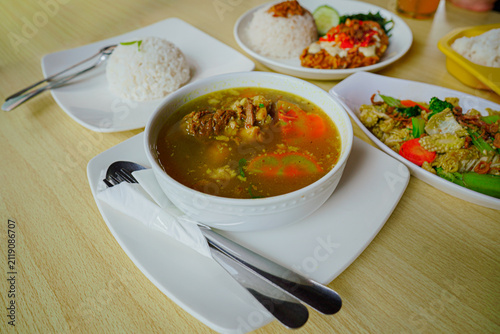 Konstfotografi Delicious Indonesian traditional beef soup served hot in a ceramic white bowl with steamed rice, fresh vegetables side dish, and cutlery on a wooden table