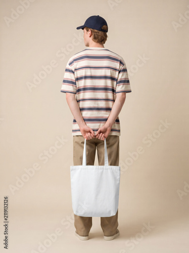 Vertical blank canvas tote bag mockup held behind the back by a young man in a striped t-shirt