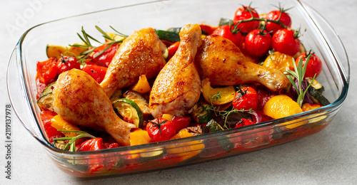 Baked chicken legs with zucchini and cherry tomatoes, garnished with parsley in glass baking dish. Grey background. Close up.