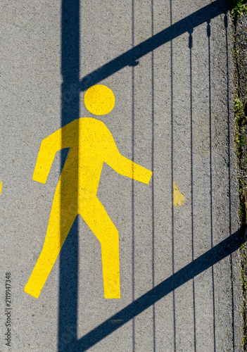 Top view of a yellow painted pedestrian sign on asphalt pavement with a metal fence shadow crossing it.