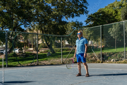 A senior man stands on a tennis court holding a racket. He is casually dressed and looks ready to play. The scene shows trees and buildings in the background under clear blue sky.