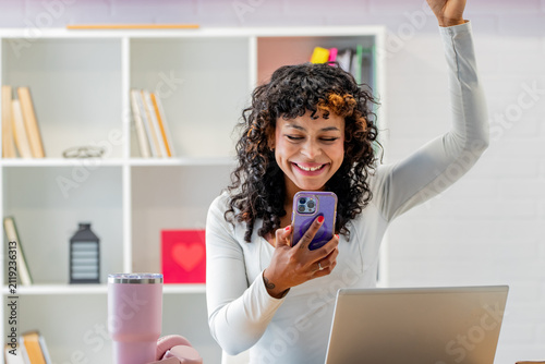 Smiling young woman with curly hair celebrating good news while looking at her smartphone, sitting at a desk with an open laptop in a bright home office