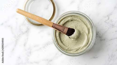 Facial clay mask in a glass jar with an applicator brush on marble