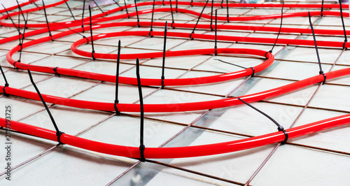 Red underfloor heating pipes installed on metal mesh grid on concrete floor, black zip ties secure pipes. Industrial construction scene with clean lines high contrast colors