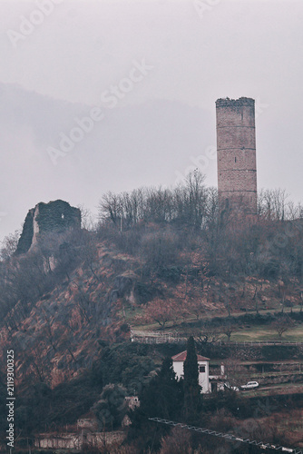 Ruined medieval stone tower rising above misty hillside with bare winter trees, collapsed fortress wall, secluded countryside house, overcast sky, quiet historic landscape