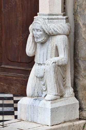 Weathered stone statue of robed human figure beside old wooden doorway with classical architectural detail, aged surface texture, historic exterior entrance and cultural heritage setting