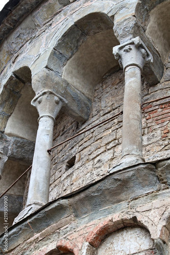 Weathered stone arches and slender columns set into aged masonry facade with rough plaster, brick textures, historic detail, and upward perspective creating strong vertical lines