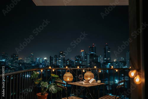 A cozy balcony at night with string lights a table two chairs potted plants lanterns a glass of red wine an open book and a smoking incense stick overlooking a vibrant city skyline