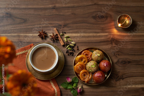 A steaming cup of Indian chai tea on a brass saucer with a plate of assorted Indian sweets jalebi ladoo gulab jamun aromatic spices a lit diya oil lamp and rosebuds