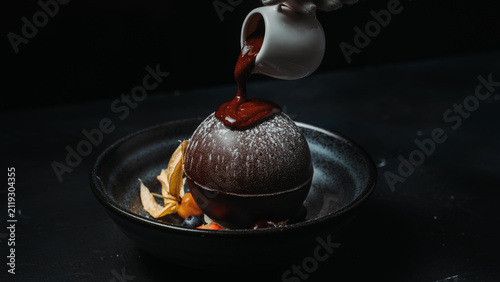 Chef pours rich chocolate sauce over an elegant chocolate sphere dessert served on a modern ceramic plate against a black background.
