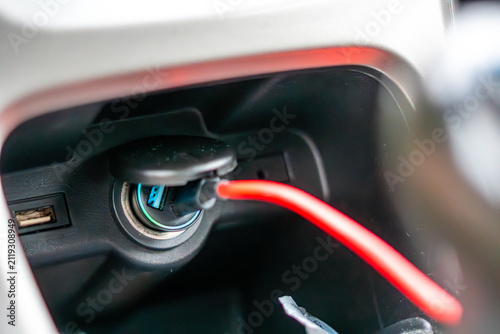 Car charger inserted into a 12V cigarette lighter socket on the vehicle center console, connected red USB cable, close-up shot of car interior, soft lighting.