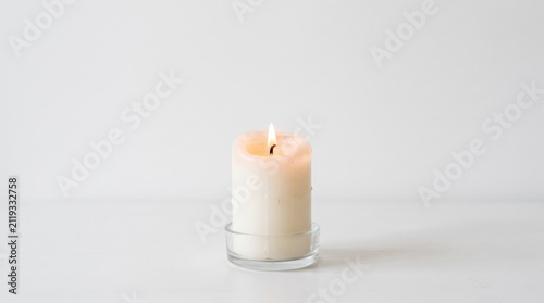 Fotografía A single white candle burning brightly on a clear glass holder against a plain w