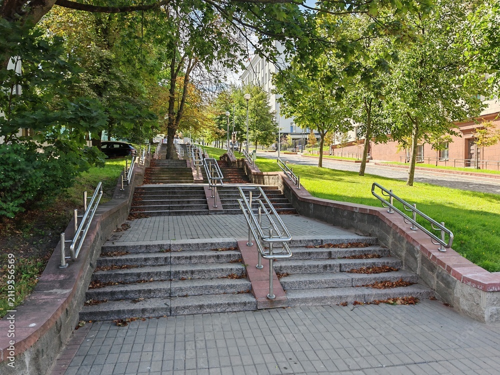 Obraz premium A staircase made of concrete tiles with metal railings has been built on the city street. There are buildings and trees nearby and the roadway is paved with paving stones. Sunny summer weather