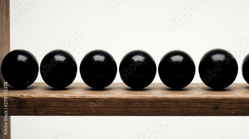 Row of matte black spheres sits on rustic light brown wooden shelf against white background. Round dark objects create contrast with grain texture of natural plank furniture. Geometric composition
