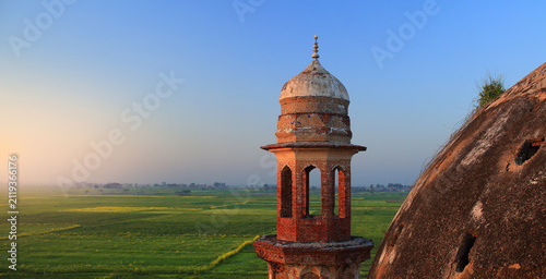 View of the historic brick minaret of the Shrine of Abdul Nabi surrounded by lush green fields under a clear sky at sunset in Kotli Maqbara, Punjab, Pakistan.
