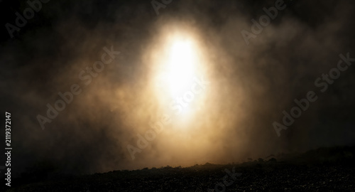 A bright, glowing oval light shines through dense fog and smoke over a dark, rocky ground, creating a dramatic, ethereal atmosphere.