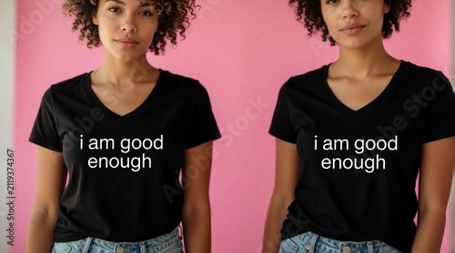 Young curly woman wears black tshirt with I good enough text slogan. Female stands against pink wall background. Person shows confidence mental health support positive message self acceptance mindset