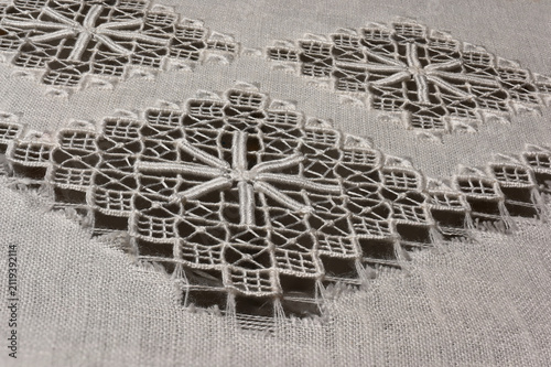 A close-up macro photograph of traditional white linen fabric featuring intricate drawn thread embroidery and decorative openwork lace.