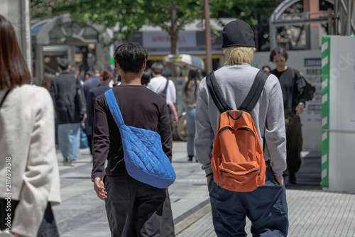 Rear view of two young adults in casual streetwear walking through a busy urban shopping district, carrying a bright blue shoulder bag and an orange backpack on a sunny day.