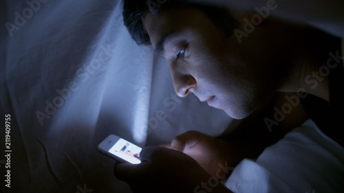 A young Indian boy / man lies under a blanket and scrolling his smartphone. 