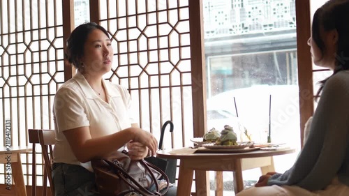 Two women converse warmly at a traditional table. Soft light filters through patterned wooden screens. One woman gestures animatedly, smiling at her companion
