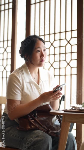 Woman sits quietly, focused on her phone. Soft light filters through wooden lattice windows. Her expression shows calm concentration. Her handbag rests nearby, untouched
