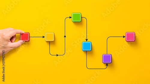 Human hand arranging colorful square blocks connected by arrows on a yellow surface. Concept of workflow optimization and logical process management.