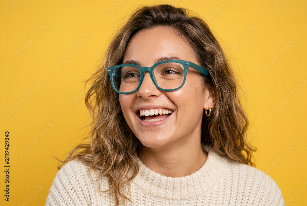 Fototapeta premium Smiling young woman wearing teal glasses and a white knitted sweater on a yellow background