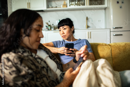 Two women using smartphones on home couch, suspicious glance