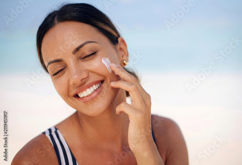 Beachside skincare moment featuring a joyful woman applying face cream or sunscreen with her eyes closed in blissful contentment