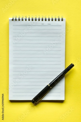 Fotografie A modern black ink pen placed diagonally across the bottom corner of an open, blank lined notepad on a vivid yellow surface